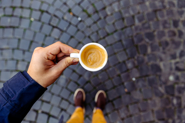 Aufnahme aus der Sicht einer Person, die auf einem Kopfsteinplaster steht, eine Tasse Kaffee in der linken Hand hält und nach unten blickt