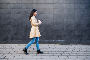 Frau mit Kaffeebecher geht entlang einer Wand und blickt lächelnd auf ihr Smartphone