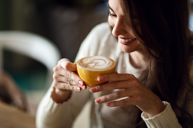 Eine lächelnde Frau blickt auf eine Tasse Kaffee mit Milchschaum, die sie mit beiden Händen vor sich hält